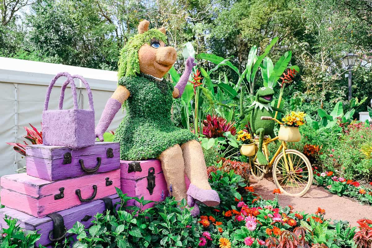 Miss Piggy sits on a topiary bundle of bags and Kermit rides a bike with a basket of flowers 