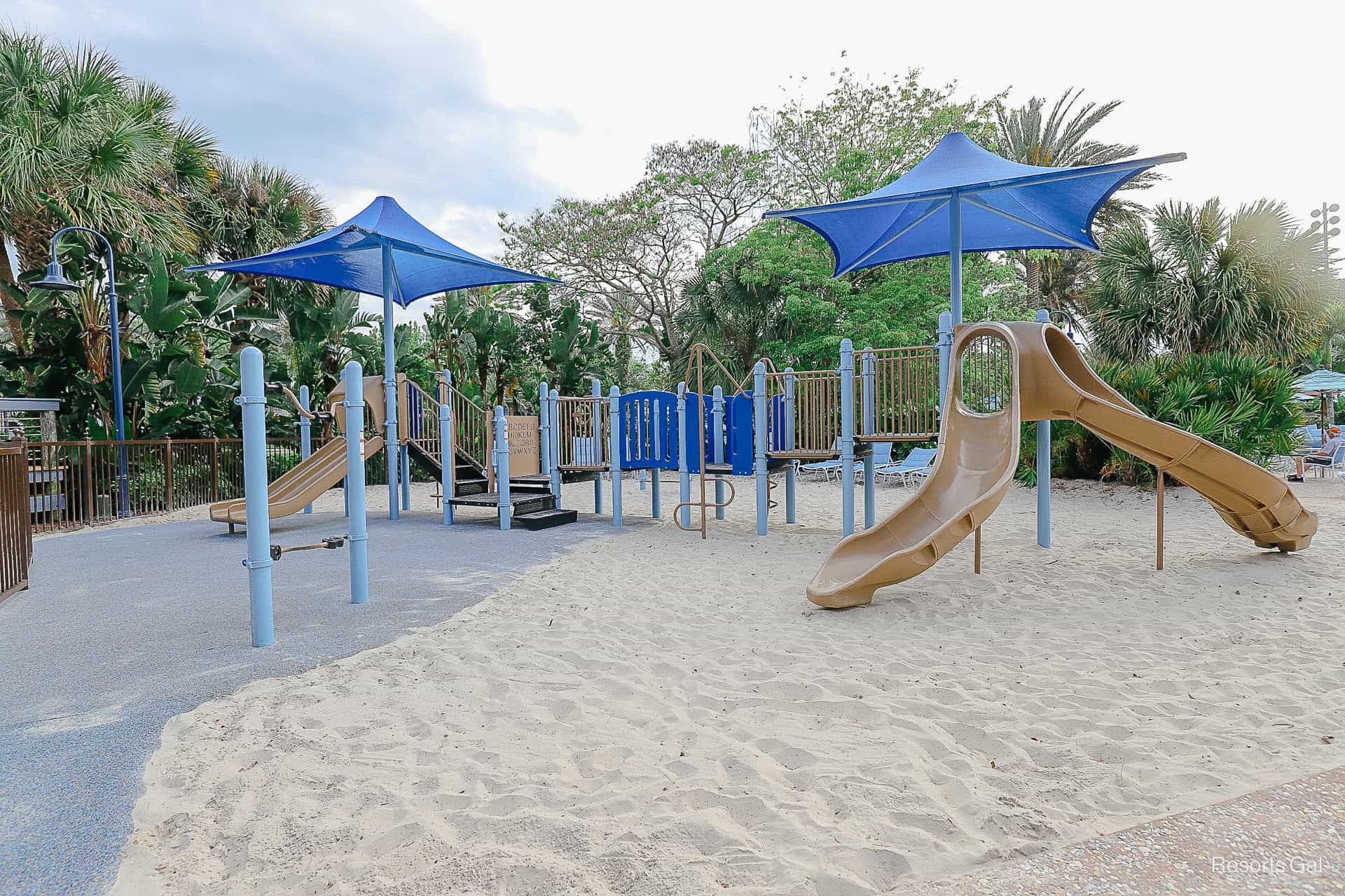 a playground with large blue umbrellas and sand and rubber surface 