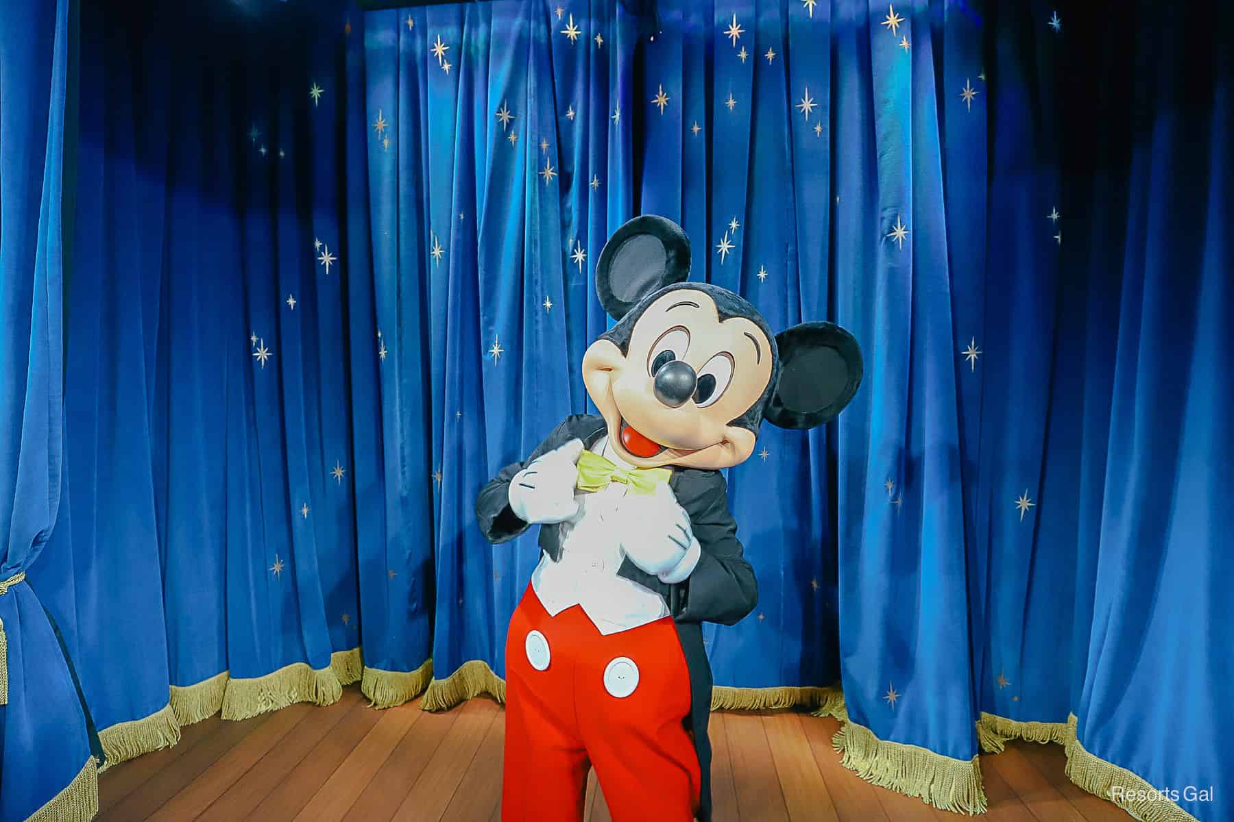 Mickey Mouse with a blue curtain behind him. He's wearing his suit and tie look. 