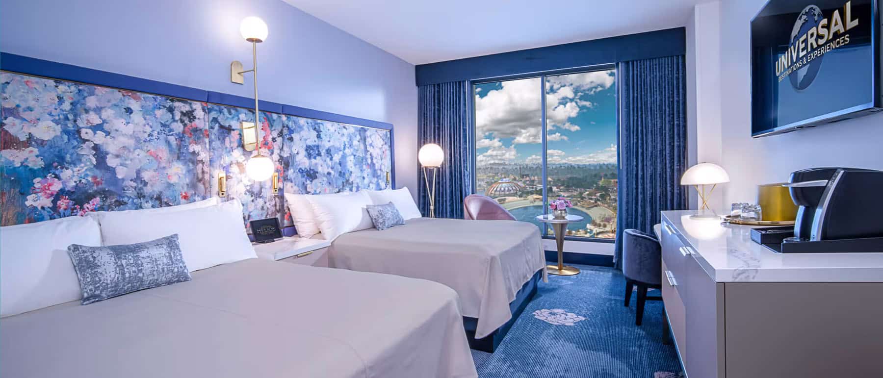 a room with light blue colors and two queen size beds that offers a view of the theme park beyond the window 